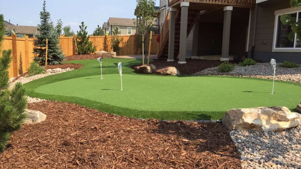 Putting green in back yard with 3 holes and flag poles in each hole. Brown mulch and rocks as well.